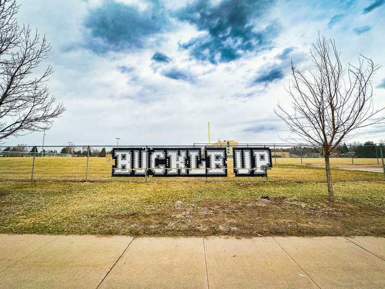 Safety first! “Buckle Up” message installed with put-in cups in chain-link fence at John F. Kennedy High School, Denver, CO