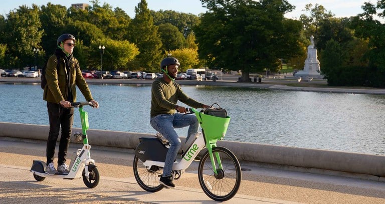 Image of two people riding an electric scooter and an electric bike through a city park. 